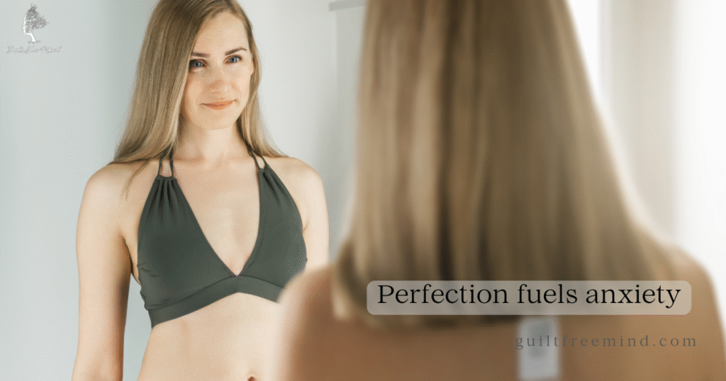 A woman in a dark green swimsuit stands in front of a mirror looking at her reflection with a gentle smile, overlay text reads “Perfection fuels anxiety” – representing how perfectionism triggers swimsuit season body anxiety.